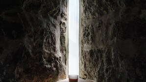 A stone-walled room with a single window at the far end, a wooden floor, and a glass of liquid on the floor. - Quinta Morazes - Casa de Campo (Ribeira Grande)