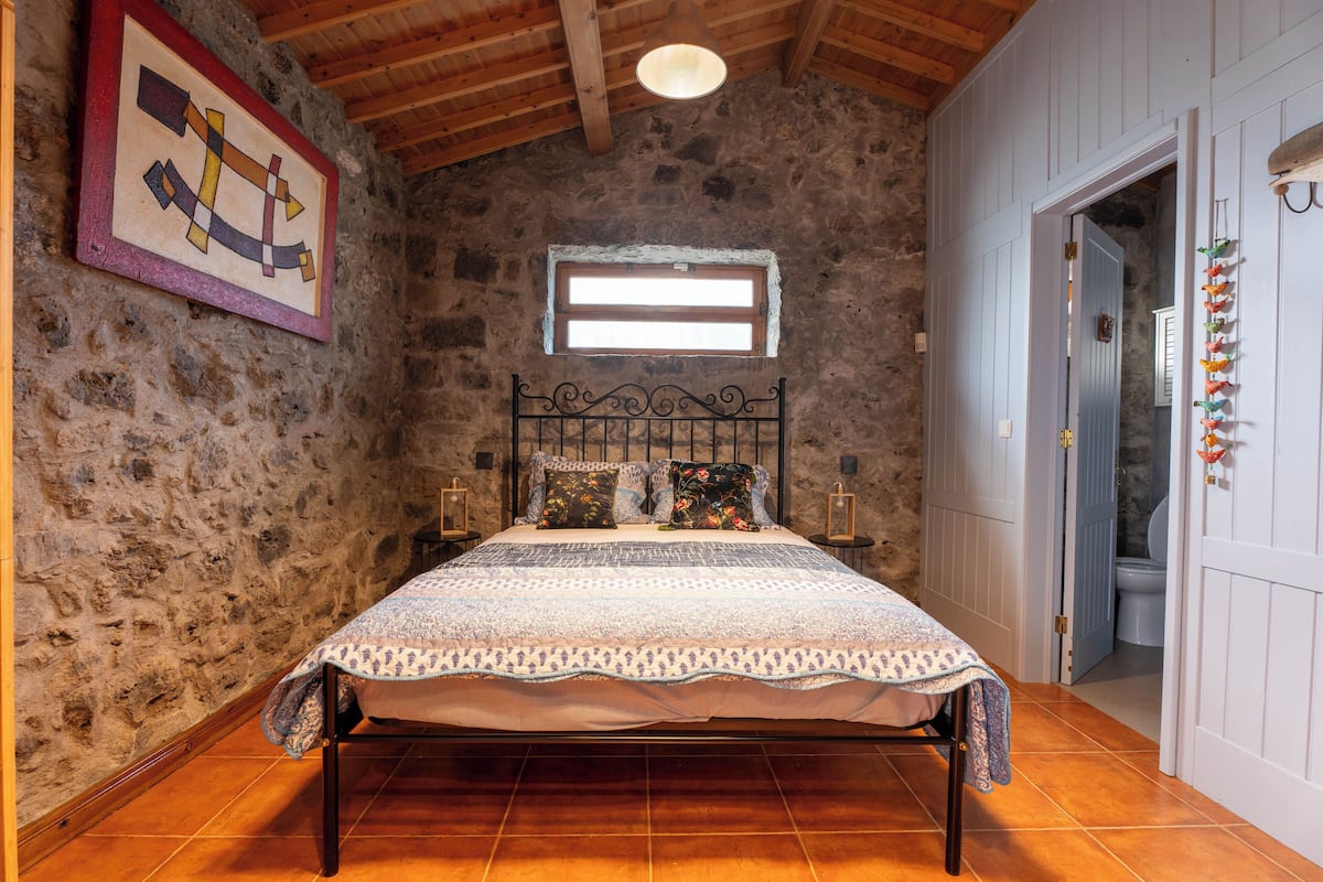 A rustic bedroom with a stone wall, a wooden ceiling, a bed with a patterned bedspread, a window with blinds, and a door leading to a bathroom.