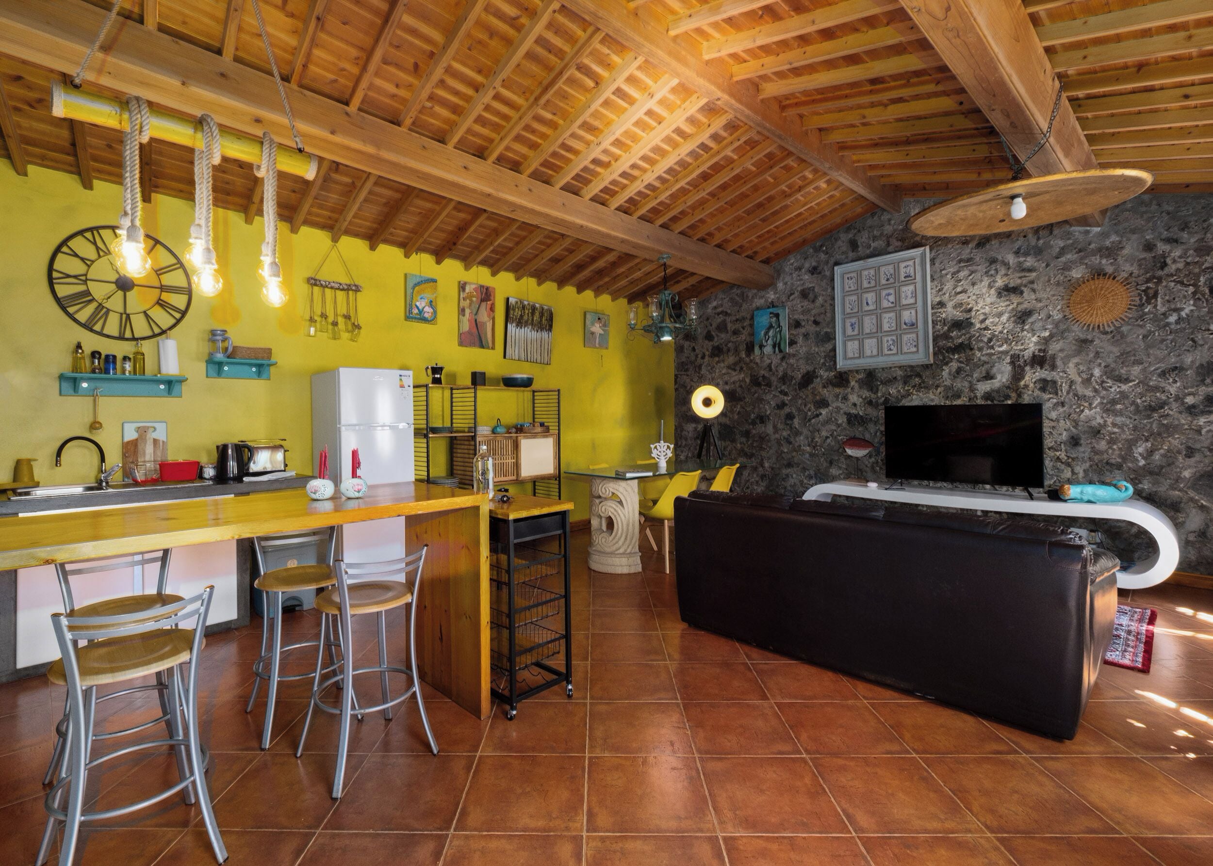 Een keuken met een houten plafond, een stenen muur en een televisie. Er staan barkrukken en een koelkast.
