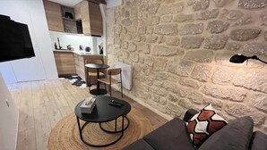 A modern living area with a stone wall, a black sofa, a round coffee table, and a kitchen in the background. - Fbg St-antoine, le Marché Aligre (Paris)