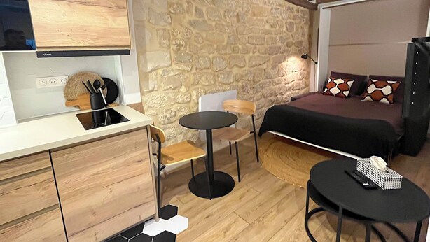 A compact room with a stone wall, a bed, a small table, a chair, and a kitchenette. - Fbg St-antoine, le Marché Aligre (Paris)