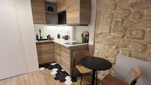 A compact kitchen with wooden cabinets, a black and white hexagonal tile floor, a small dining table with two chairs, and a stone wall. - Fbg St-antoine, le Marché Aligre (Paris)