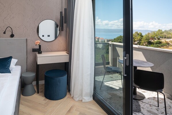 A modern hotel room with a bed, bedside table, mirror, and a view of the sea through a sliding glass door.