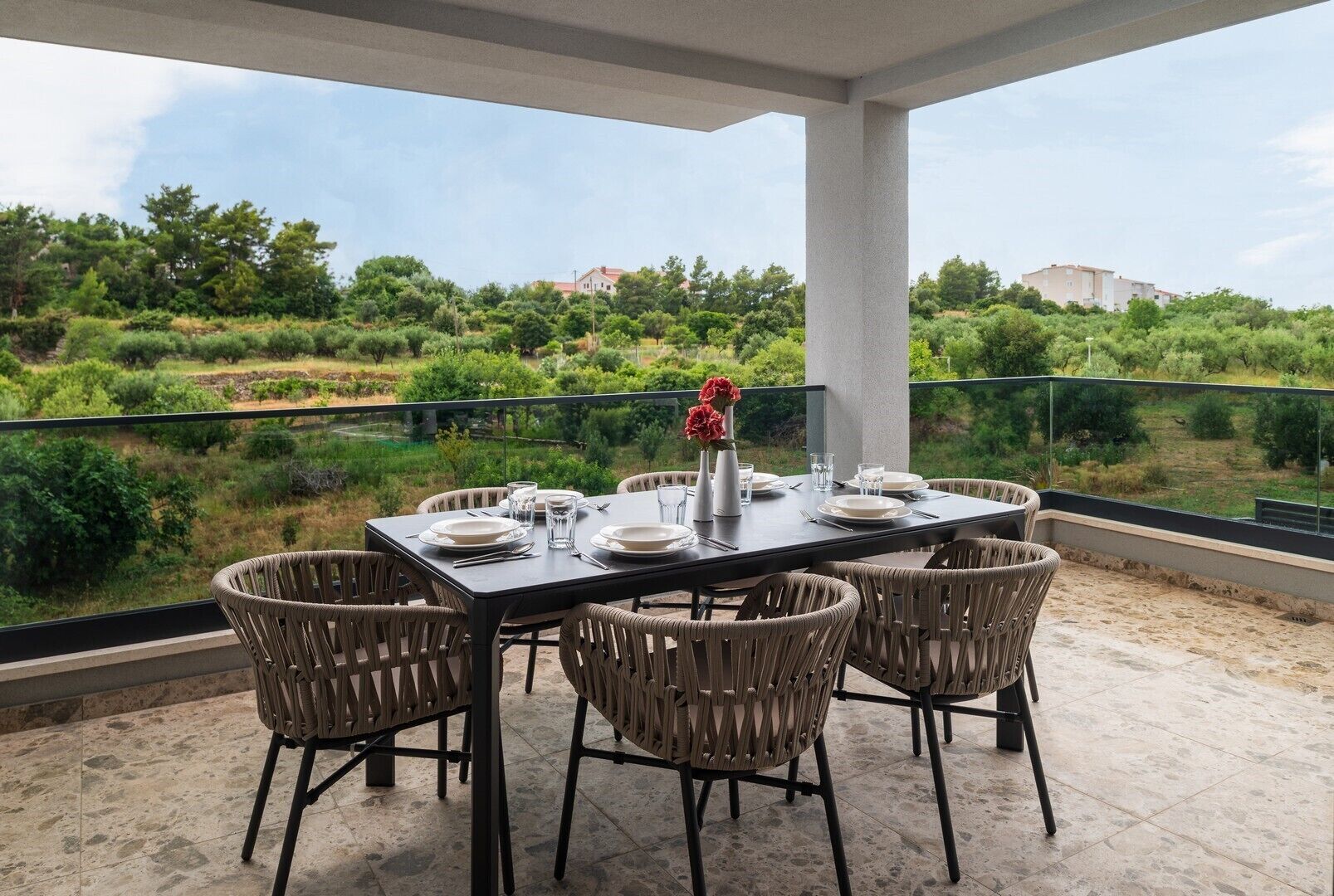 A covered outdoor dining area with a table set for six, surrounded by wicker chairs, overlooking a lush landscape with trees and buildings in the distance.
