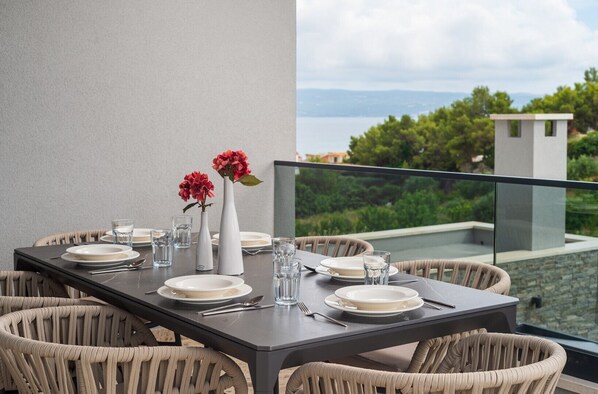 A dining table set with plates, glasses, and cutlery, surrounded by wicker chairs, with a view of greenery and a building in the background.