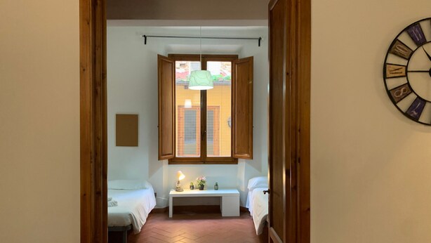 A narrow hallway leading to a bedroom with two beds, a small bedside table with a lamp, and a window with blinds. - Sdrucciolo dei Pitti First Floor (Florence)