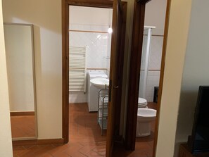 Ένας στενός διάδρομος οδηγεί σε ένα μπάνιο που περιλαμβάνει τουαλέτα, νιπτήρα και κρεμάστρα για πετσέτες.