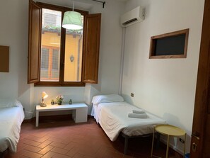 Ein kleines, sauberes Zimmer mit zwei Betten, einer Bank, einem kleinen Tisch und einem Fenster mit Jalousien.