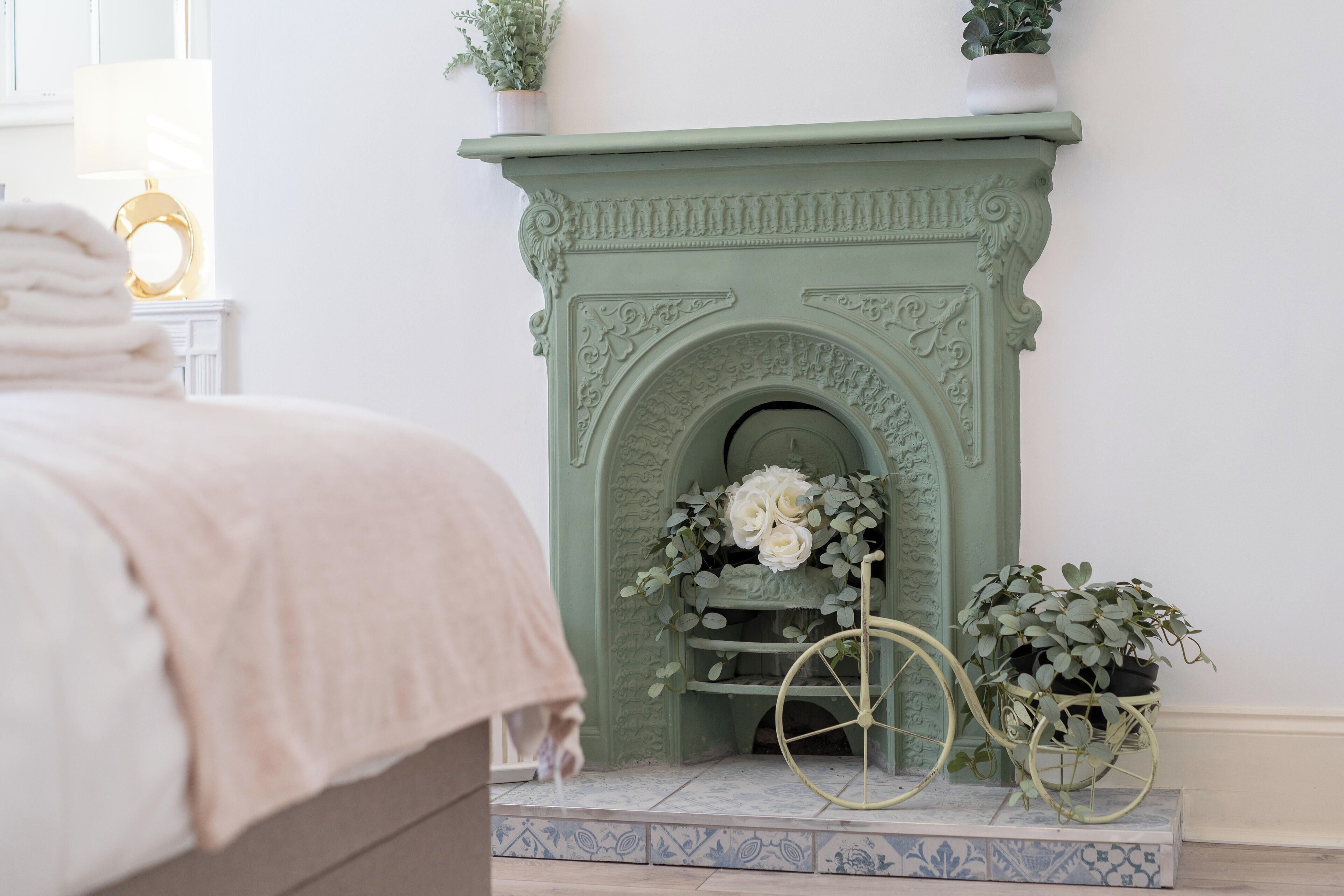 A bedroom with a green fireplace, a bed with a beige blanket, and a small potted plant on the windowsill.
