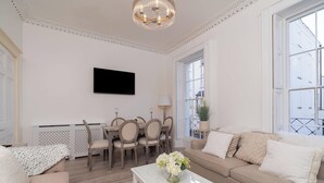 Ein hell erleuchtetes Wohnzimmer mit einer Couch, einem Couchtisch mit Blumen, einem Essbereich mit Stühlen und Tisch sowie einem großen Fenster mit Blick auf Gebäude.