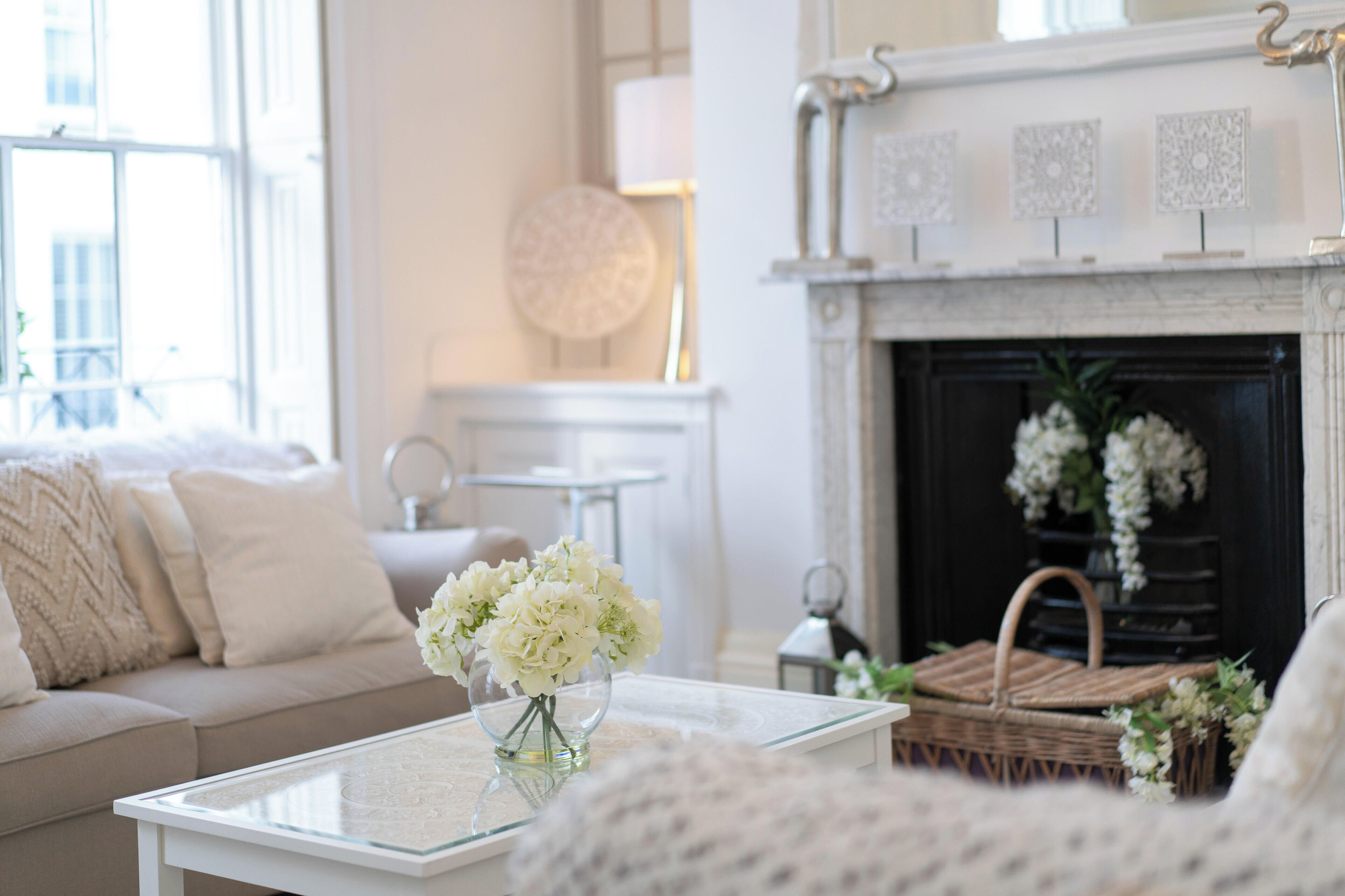 Un salon avec un canapé blanc, une table basse en verre ornée d’un vase de fleurs, une cheminée décorée de carreaux et un panier en osier.