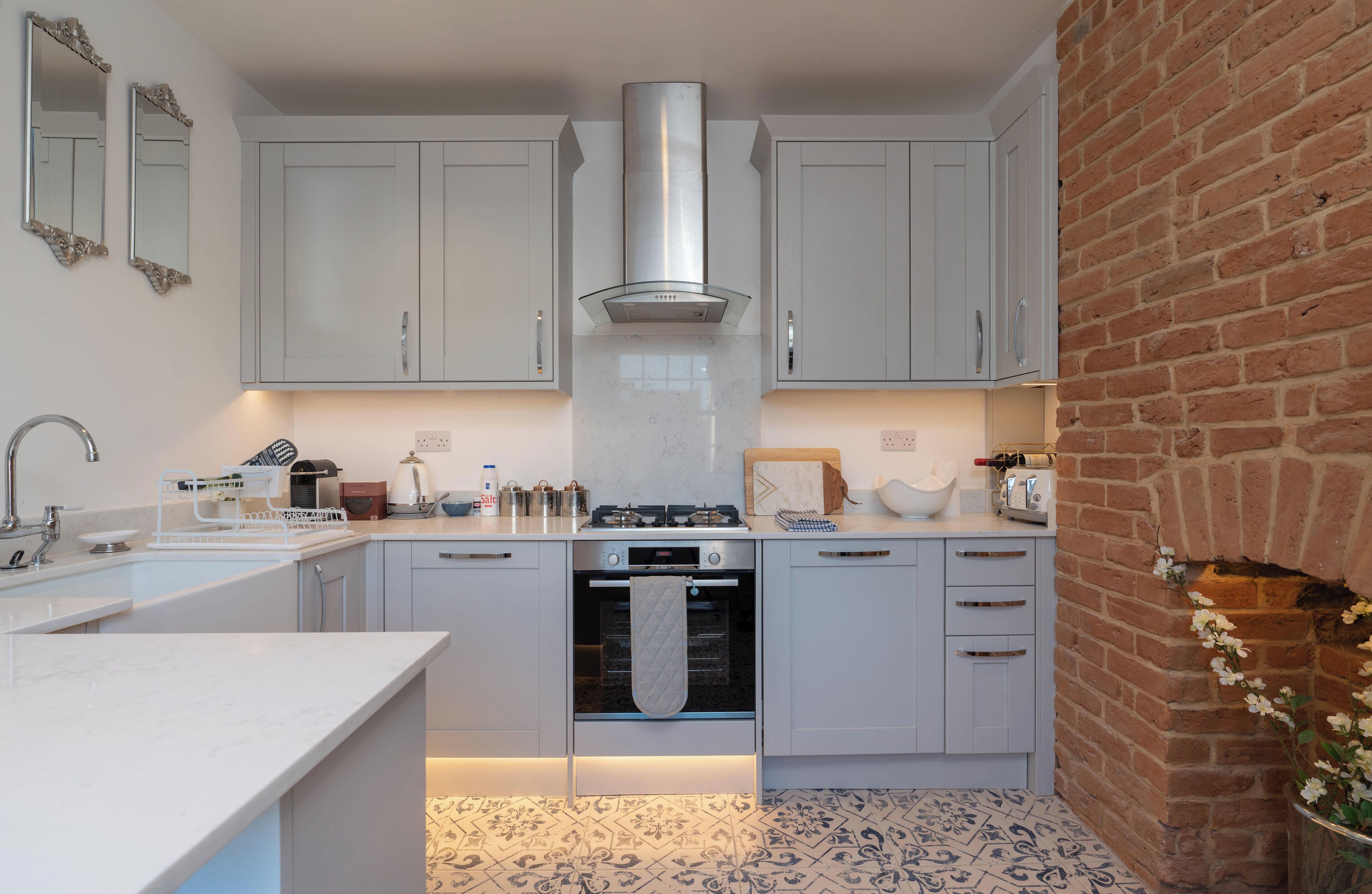 A modern kitchen with white cabinets, a stainless steel range hood, and a brick wall.