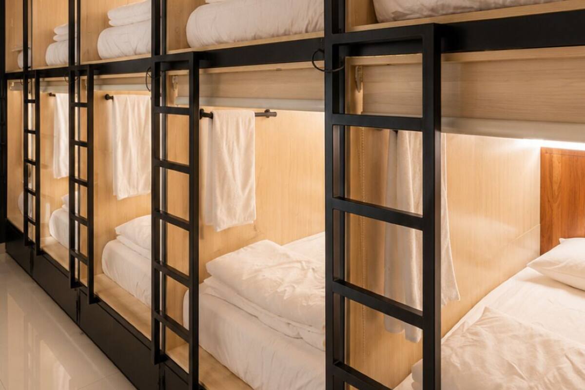A row of bunk beds with wooden frames and white bedding, a ladder, and a shelf with bottles.