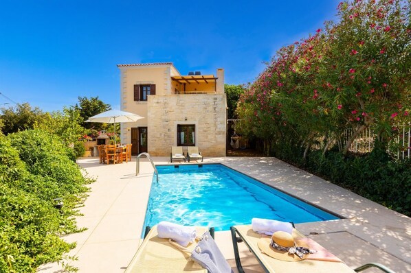 A swimming pool with lounge chairs and a table, surrounded by greenery and a stone building.