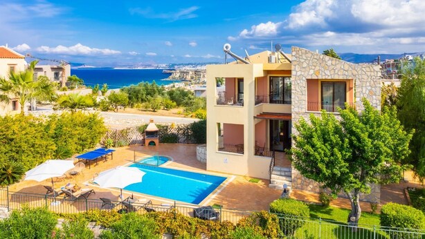 Un edificio de dos pisos con piscina, mobiliario exterior y vista al mar.