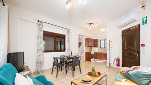 Una sala de estar con un sofá, una mesa de centro con fruta, una zona de comedor con mesa y sillas, una cocina al fondo y una puerta con extintor.