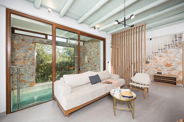 A modern living room with a sofa, a coffee table, and a chair. There is a staircase with a wooden railing and a stone wall. - Villa Nineta - Three Bedroom Villa, Sleeps 6 (Tavronitis)
