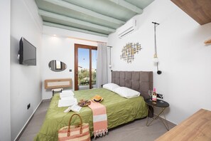 A modern hotel room with a green bedspread, a wooden headboard, a flat-screen TV, a bedside table with a bottle and glass, and a view of the outdoors through a sliding door. - Villa Nineta - Three Bedroom Villa, Sleeps 6 (Tavronitis)