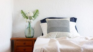 Een net opgemaakt bed met een witte deken, een nachtkastje met een bloevas, en een wandlamp-schakelaar.