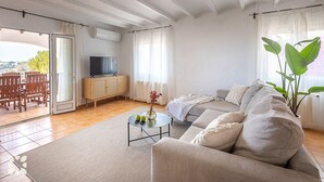 Een ruime woonkamer met een bank, een salontafel, een televisie op een kast en uitzicht op de omgeving.