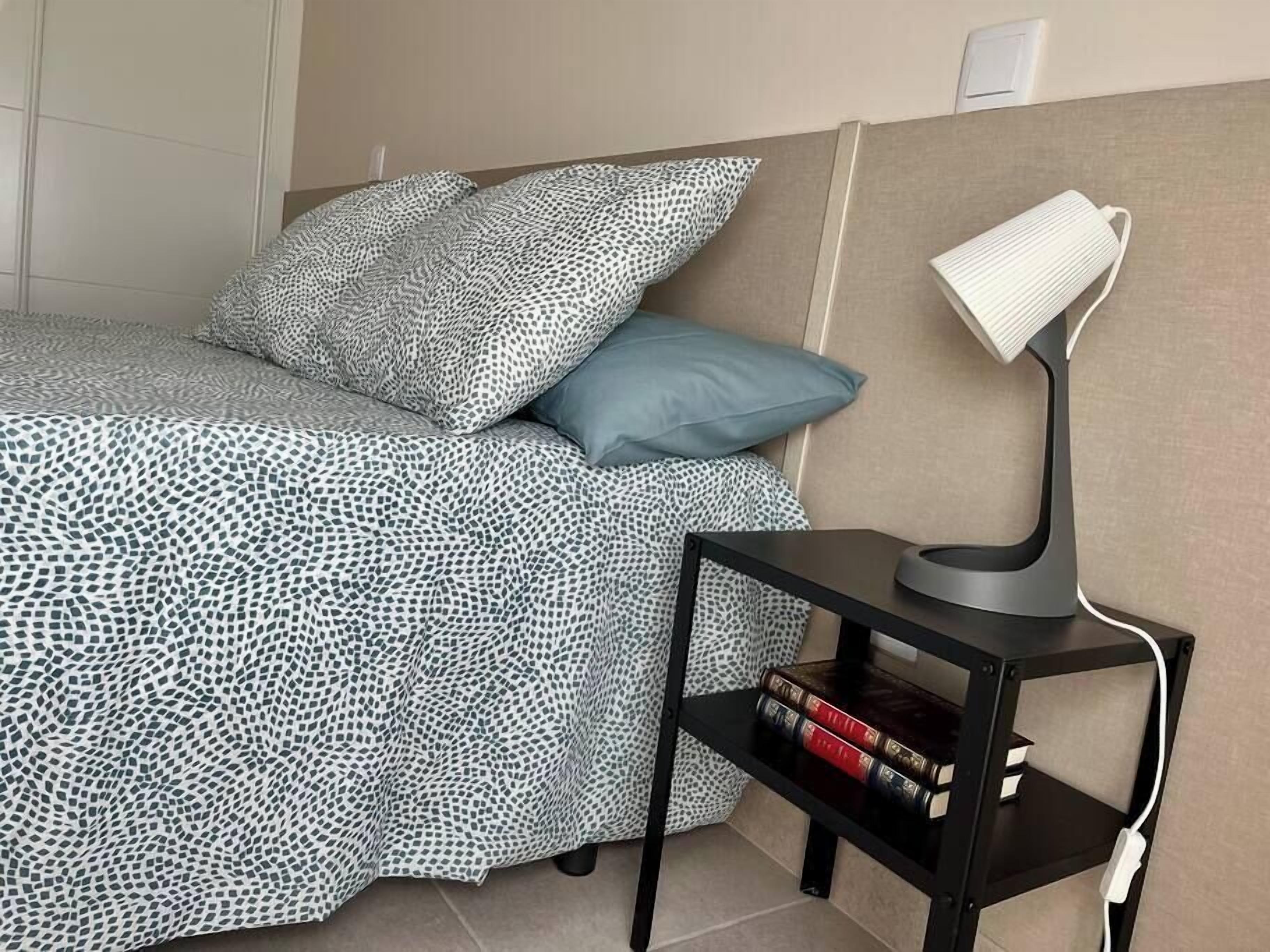 Een net opgemaakt bed met beddengoed met een patroon, een bedlamp en een klein plankje met boeken.