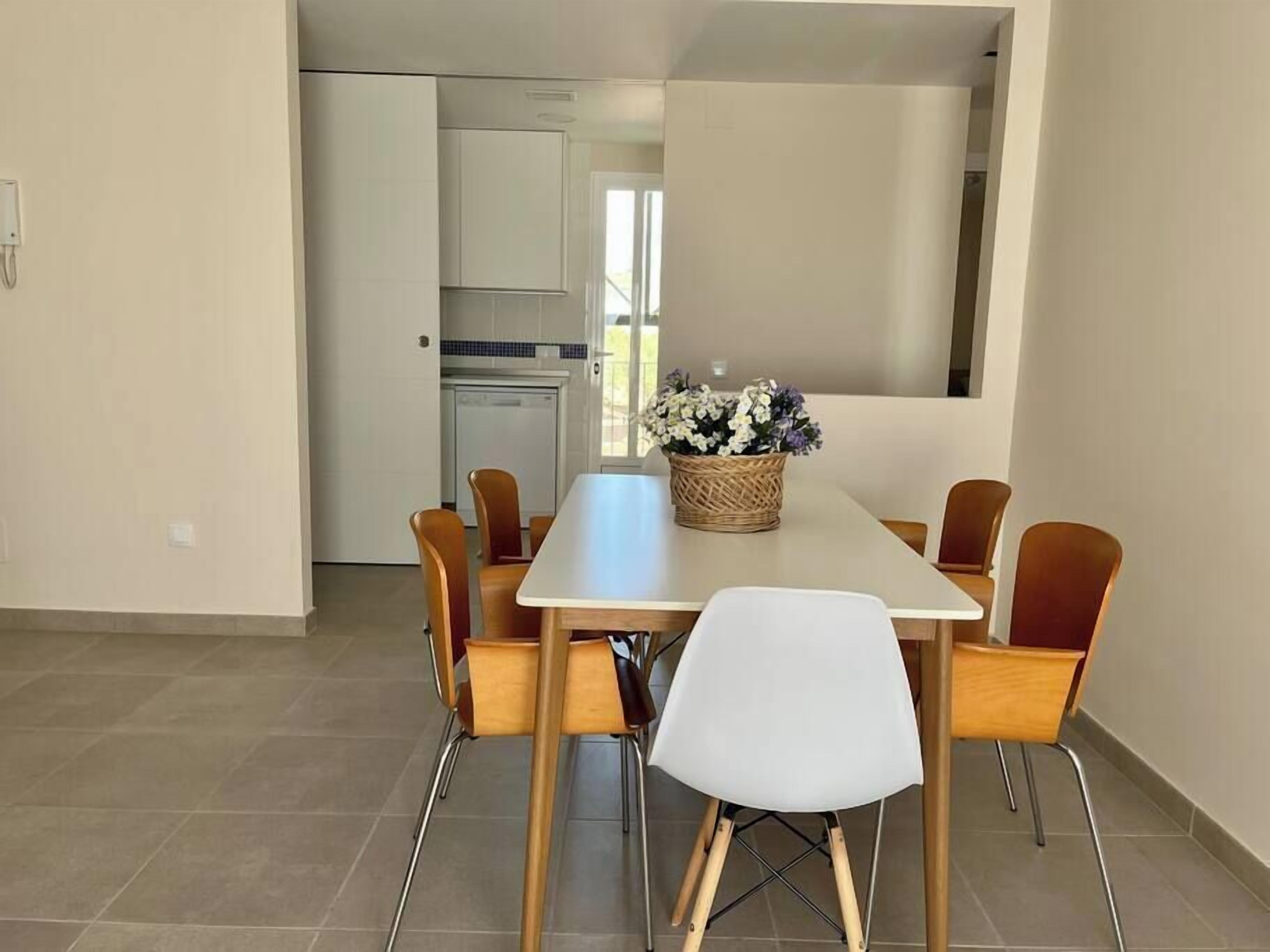 Een eetruimte met een lange tafel, zes houten stoelen en een witte stoel in het midden. Een rieten mand met bloemen staat op de tafel.