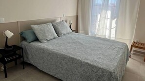 Un dormitorio con cama, dos almohadas, una mesita de noche con lámpara y una ventana con cortinas traslúcidas.