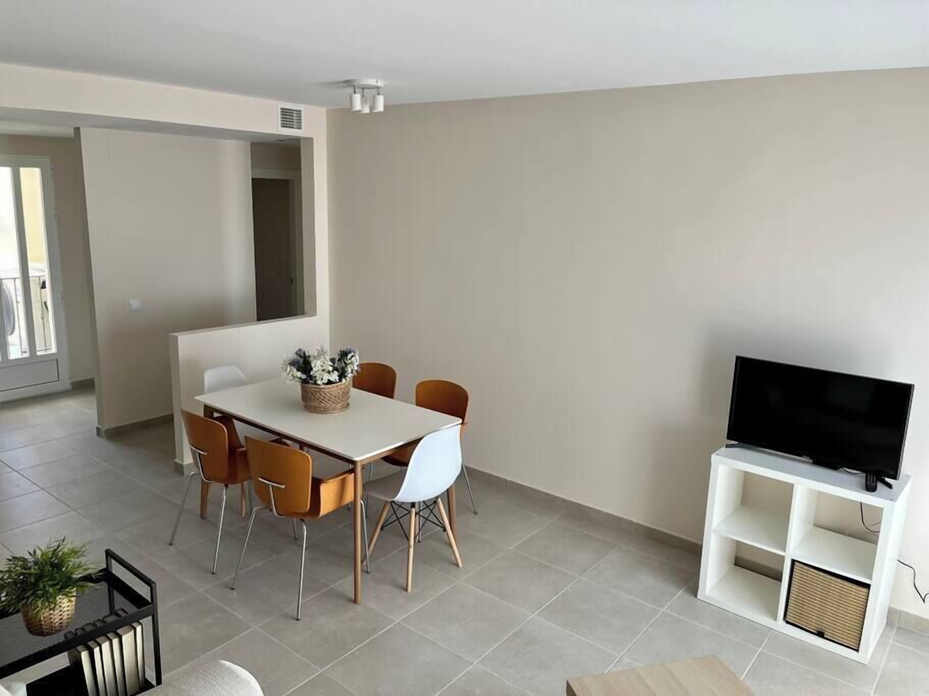 A modern living room with a dining area, a flat-screen TV on a white stand, and a large window providing natural light.