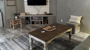 A living room with a flat-screen TV mounted on a white wall, a wooden coffee table, a grey sofa, a patterned rug, and a bookshelf filled with books and decorative items. - Modern 2-bdrm/2-bath mobile home, near Wildcat ATV Park, riders welcome! Wi-Fi! (Livingston)