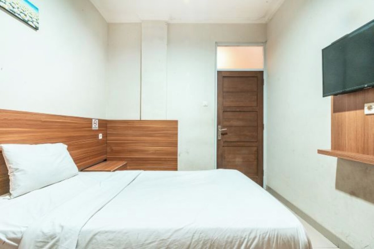 A bedroom with a wooden headboard, a bed with white bedding, a television mounted on the wall, and a door leading to another room.