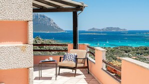 A balcony with a table and chair, overlooking a coastal view with boats and a mountain in the distance.