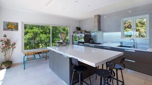 A modern kitchen with a large island, built-in ovens, a stainless steel range hood, and a window overlooking greenery.