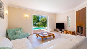 A spacious living room with a large sofa, a coffee table, and a television. There is a sliding glass door leading to a pool area.