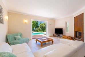 Un salón amplio con un sofá grande, una mesa de centro y un televisor. Hay una puerta corrediza de vidrio que da a una zona de piscina.
