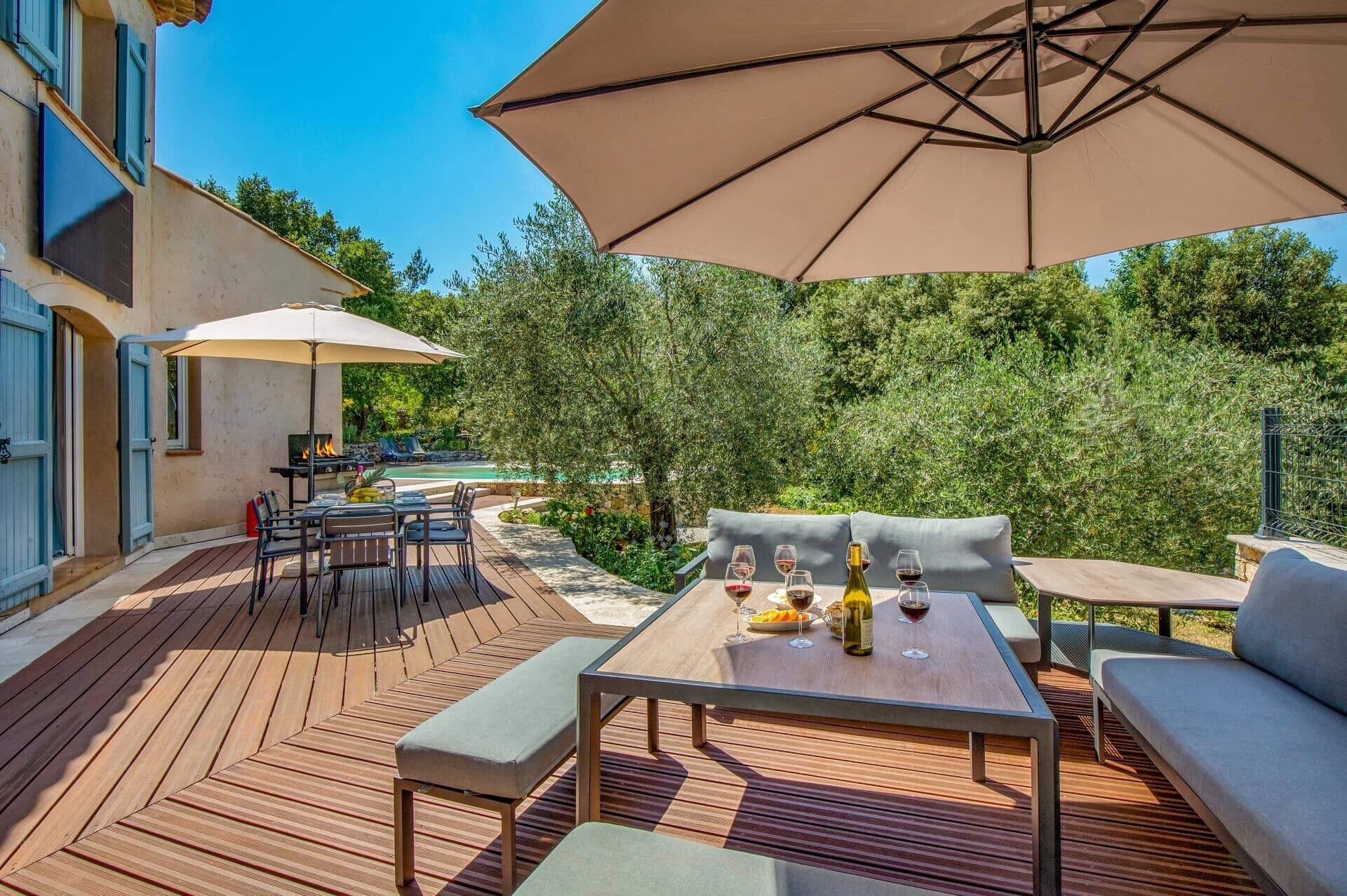 An outdoor patio with a wooden deck, a table set for dining, and a large umbrella providing shade.