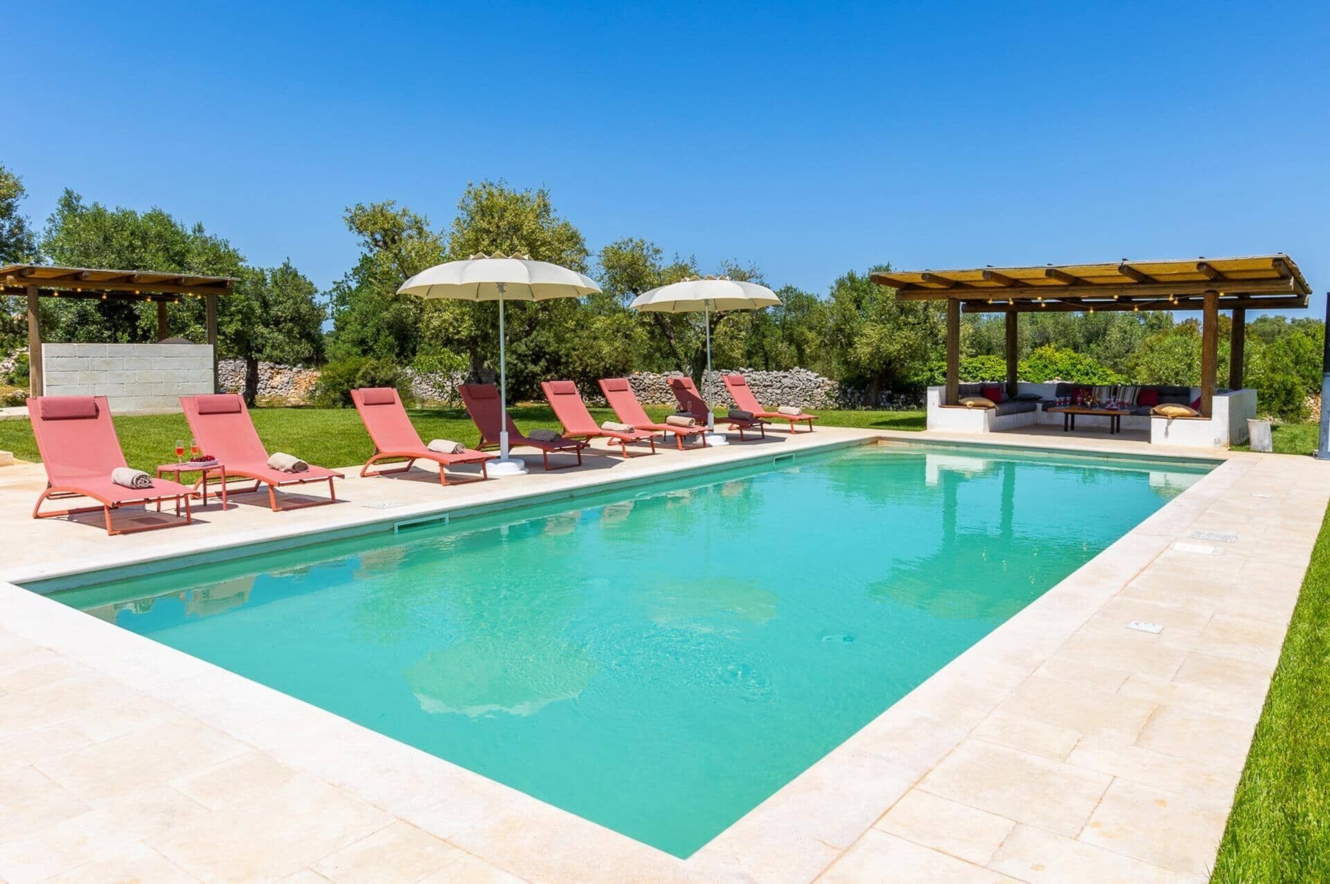 A swimming pool with red lounge chairs and umbrellas, a pergola with string lights, and a stone wall in the background.