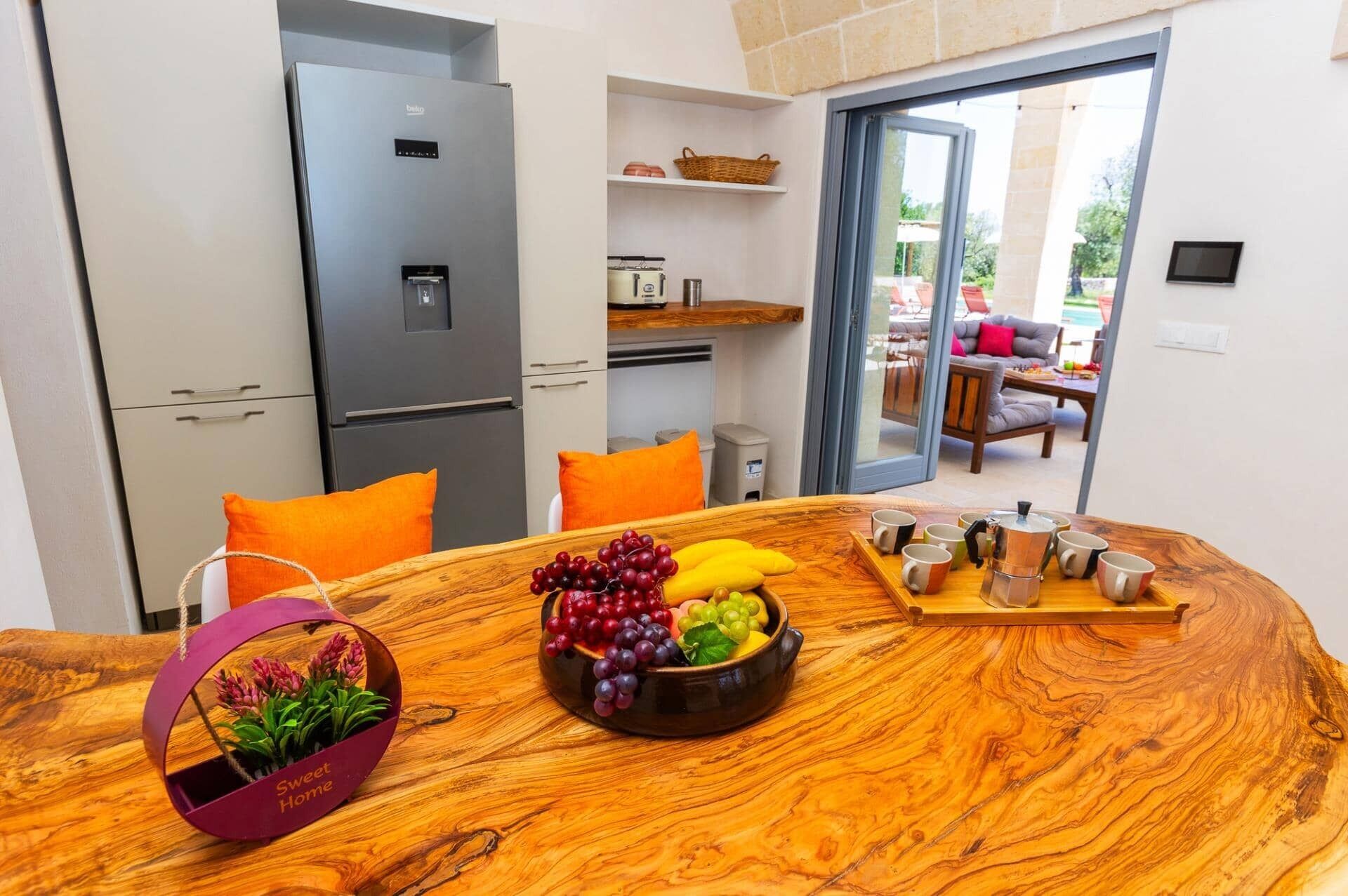 A modern kitchen with a built-in refrigerator, a dining table with fruit and coffee, and a view of the outdoor seating area.