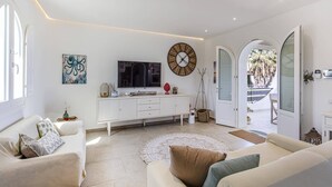 Un salón amplio con un sofá blanco, un televisor de pantalla plana, un reloj decorativo y un ventanal grande.