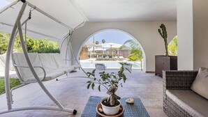Un patio iluminado por el sol con una silla mecedora, una zona de estar con cojines, una planta en maceta y vistas a una piscina y edificios al fondo.