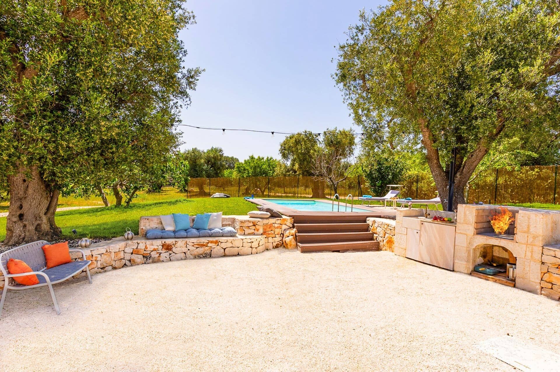 Un patio exterior con un banco de piedra, una parrilla y una piscina al fondo.