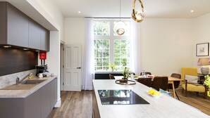 Une cuisine moderne avec un îlot central, un coin repas agrémenté d’une chaise jaune, et une vue sur la verdure grâce à de grandes fenêtres.