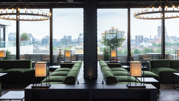 A modern lounge area with green sofas, tables, and lamps, featuring large windows with a city view. - Stylish New Studio Apartment - Battersea (London)