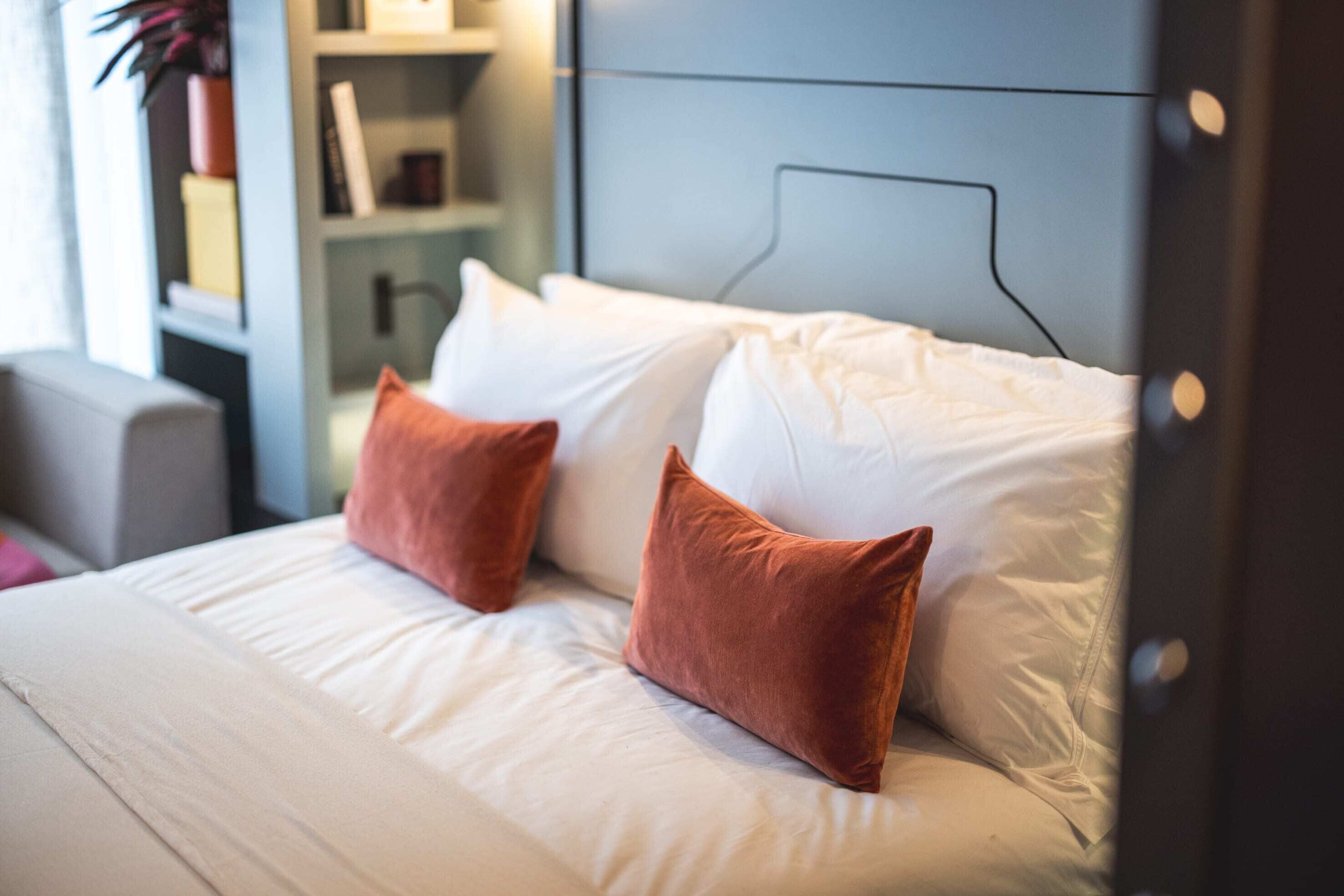 Uma cama bem arrumada com lençóis brancos e duas almofadas laranja, uma cabeceira moderna e uma estante de livros ao fundo.