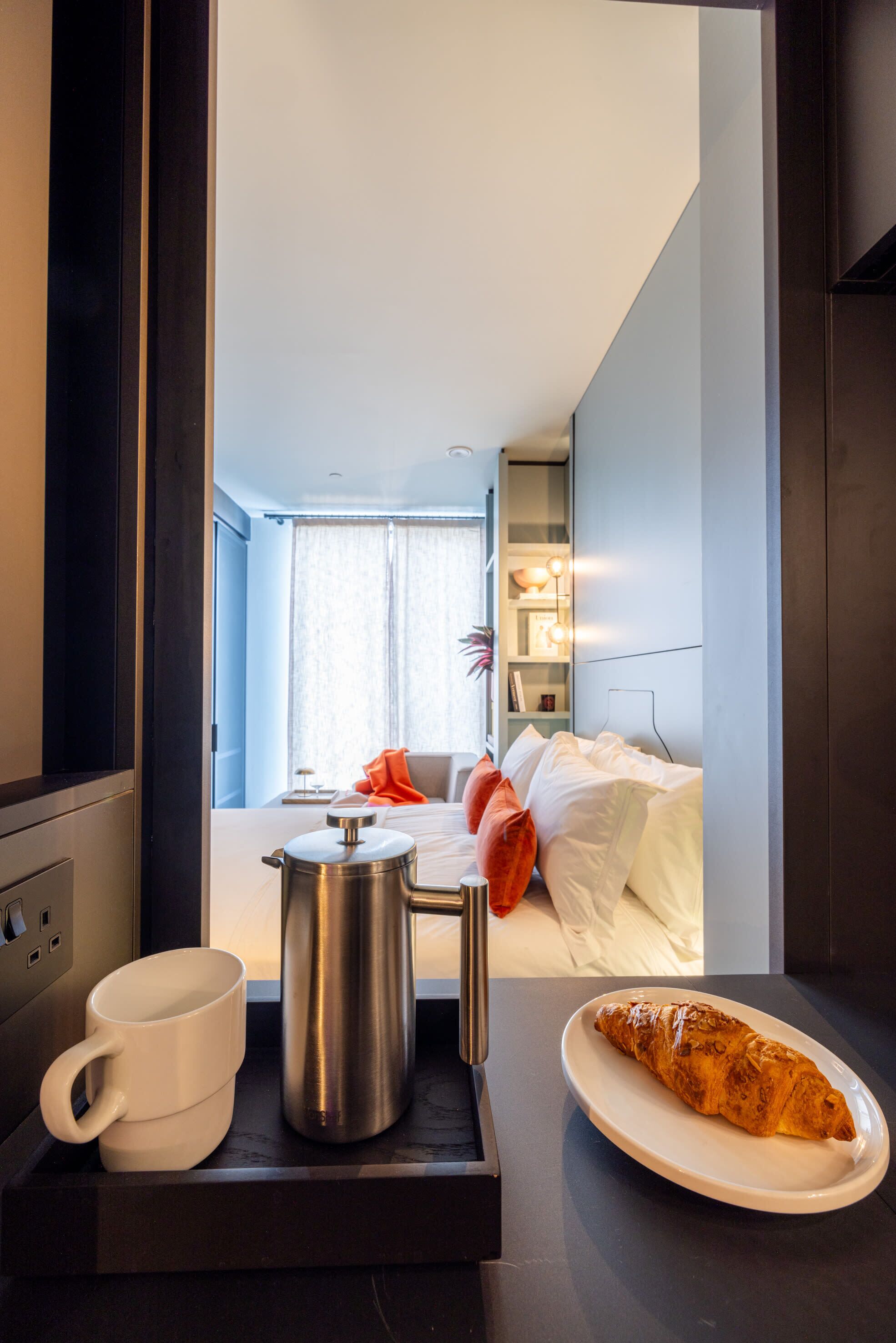 A hotel room with a bed, a bedside table with a coffee pot, a mug, and a plate with croissant.