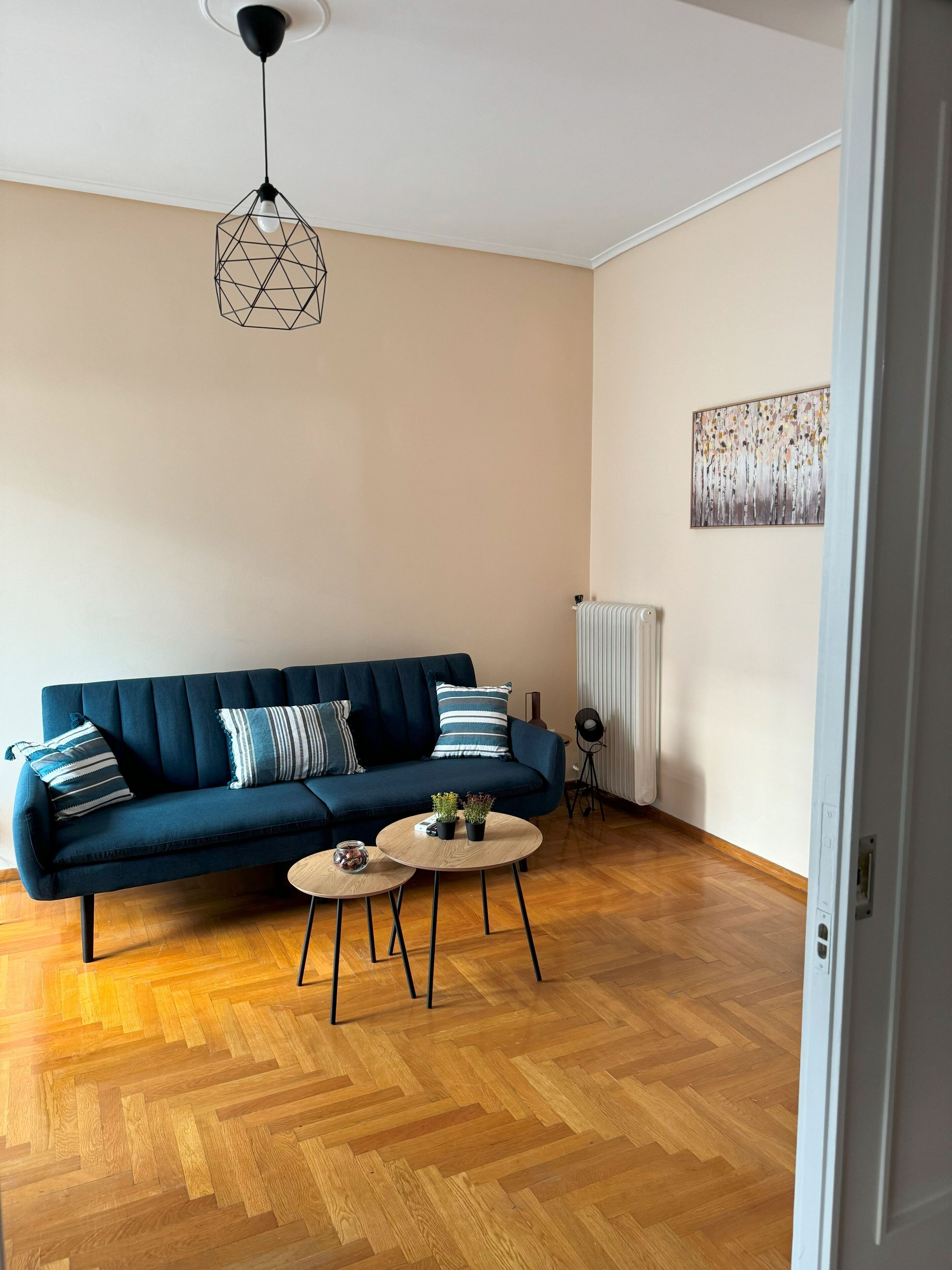Una sala de estar con un sofá azul oscuro, dos mesitas de centro de madera redondas, una lámpara colgante geométrica y un radiador.