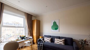 Ein modernes Wohnzimmer mit einem dunkelblauen Sofa, einem Esstisch mit Stühlen, einem großen Fenster mit Vorhängen und einem grünen, abstrakten Gemälde an der Wand.