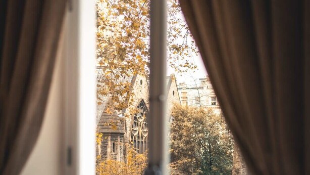 A room with a large window, curtains, and a view of a building with trees.