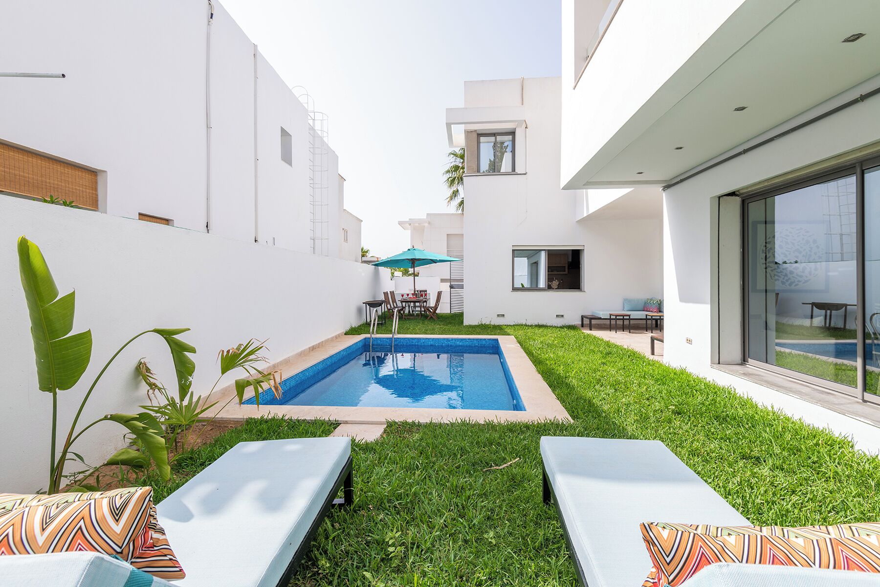 A modern outdoor pool area with a rectangular pool, two lounge chairs, and a small table with an umbrella.