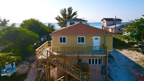 Un edificio de dos pisos con balcón, cerca de una playa donde se ve un barco amarrado.