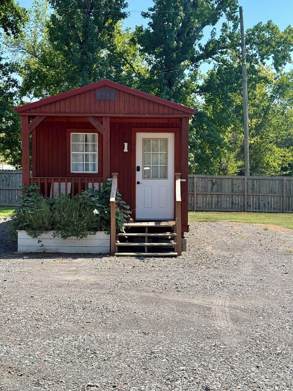 A small red wooden cabin with a white door and windows, surrounded by gravel and a wooden fence. - Cheerful cabin with KING bed and WiFi, AC in charming Plainview (Plainview)